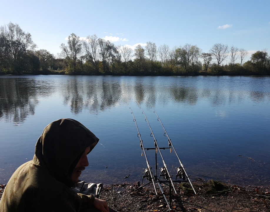 Carp angler wearing a thick hooded jacket sitting on the bank of a lake fishing with three rods on bite alarms during winter.