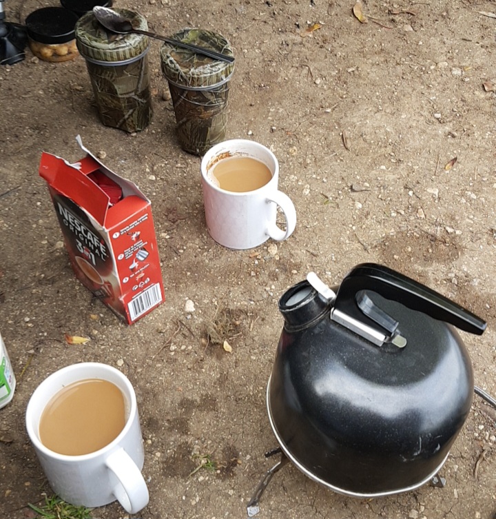 Winter carp fishing ground setup with a black kettle, Nescafe sachets, and two mugs of hot tea beside insulated thermal tumblers.